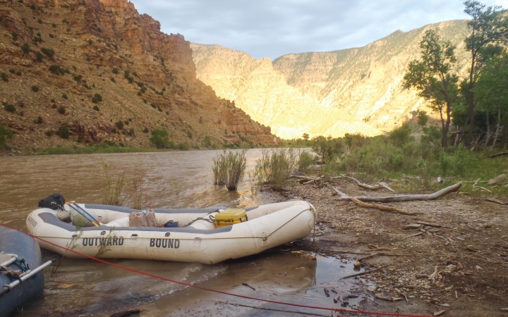 An empty raft rests on the shore of a river. In the background there are tall canyon walls.